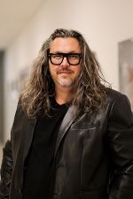 Person mit langen, grauen Haaren und Brille trägt eine dunkle Jacke. Porträtansicht.