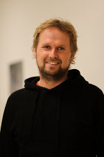 Person mit kurzen, blonden Haaren und Bart, lächelnd in die Kamera.