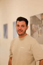 Person mit dunklem Haar und Bart lächelt in die Kamera. Im Hintergrund eine Wand mit Kunstwerken.