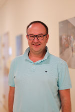 Lächelnde Person steht vor hellem Hintergrund und trägt ein Poloshirt.