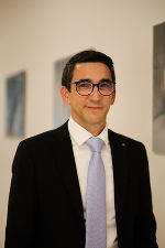 Person mit dunklem Haar und Brille, gekleidet in dunklen Anzug und hellblauem Hemd.