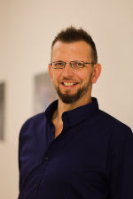 Person mit Brille und Bart lächelt in die Kamera. Dunkelblaues Hemd, hellgrauer Hintergrund.